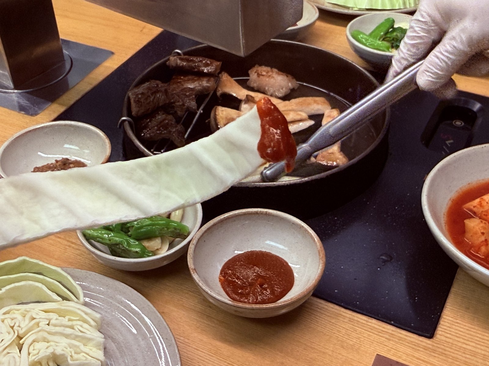 Daedo staff in white glove spreading fresh red gochujang stripe across a cabbage leaf with tongs over the grill