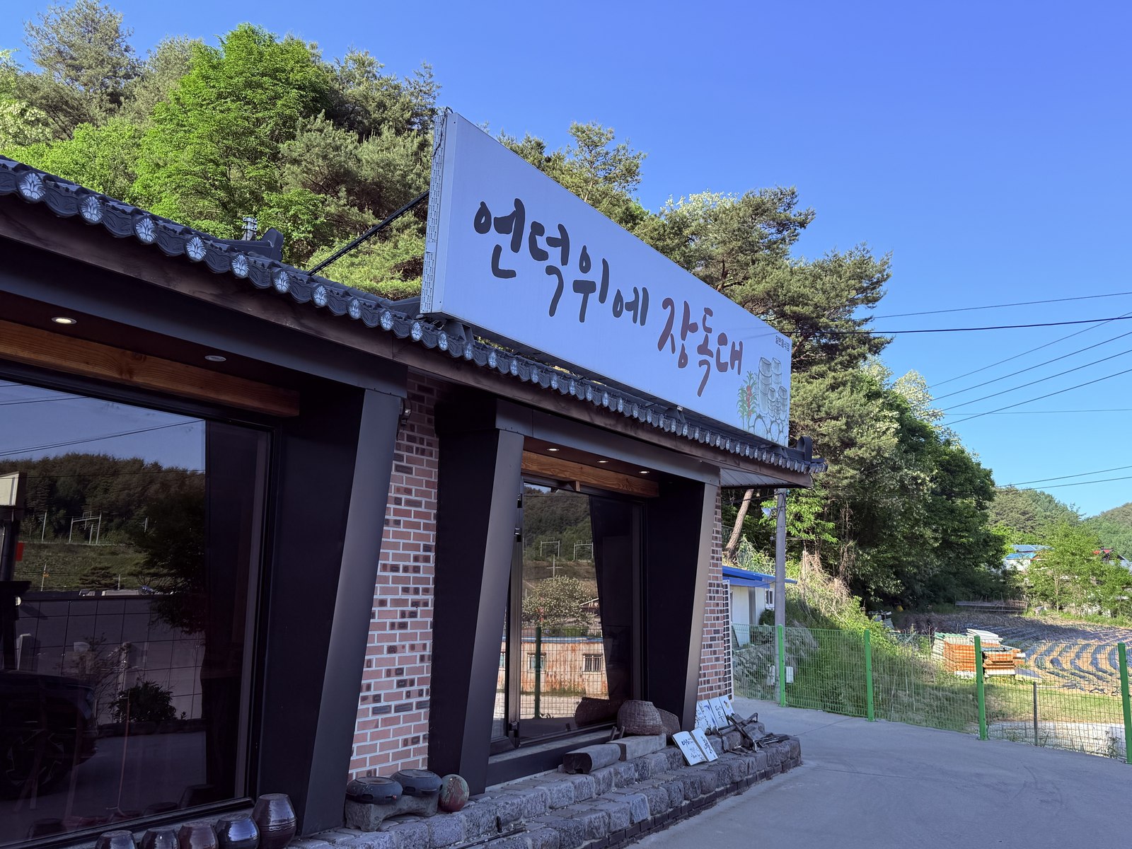 Eondeokwie Jangdokdae signage - large white sign with handwritten Korean calligraphy 언덕위에 장독대, traditional curved tile roof, mountain forest in background