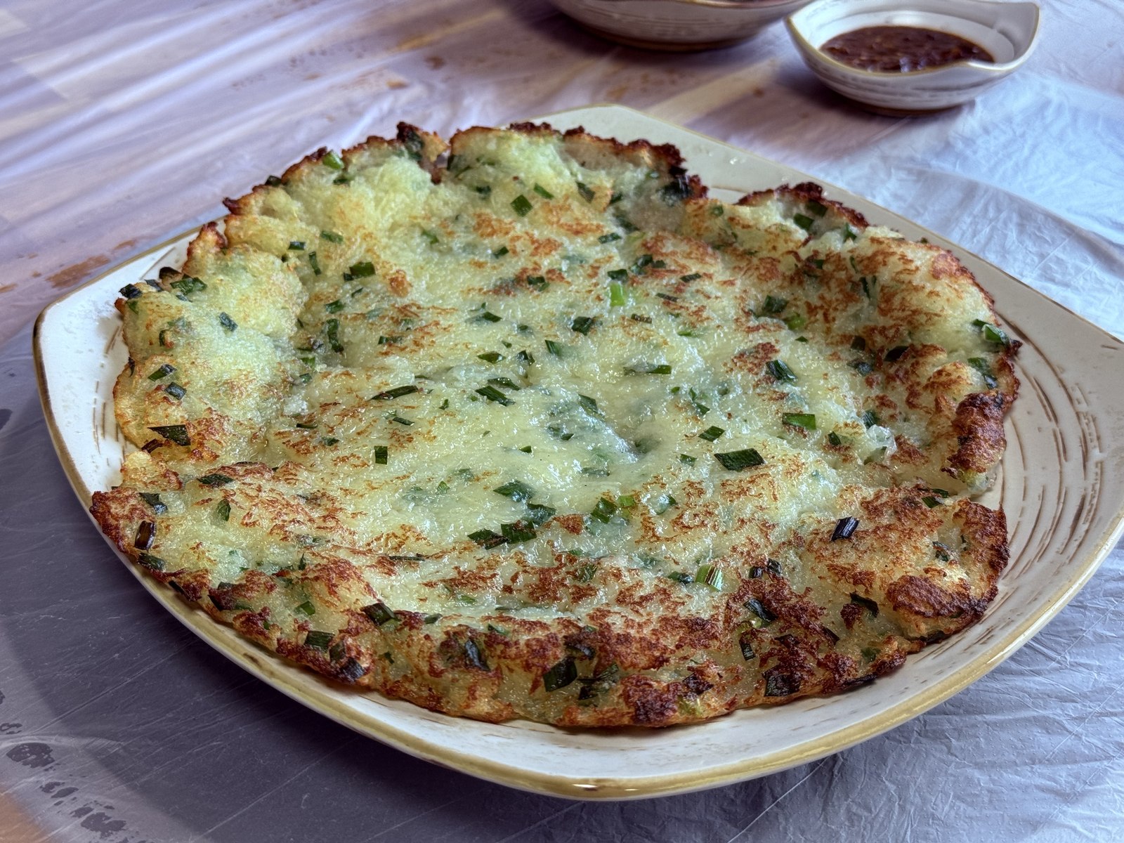 Close-up of Eondeokwie Jangdokdae potato-jeon - large round Korean potato pancake with crisp golden lace edges and translucent half-set center, chopped green onion flecks, on a rustic ceramic plate