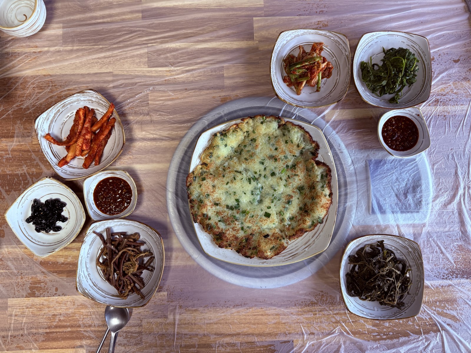 Top-down view of the Eondeokwie Jangdokdae table - large round potato-jeon in the center, eight small bowls of side dishes arranged around it including anchovies, mountain greens, kimchi, and chili paste