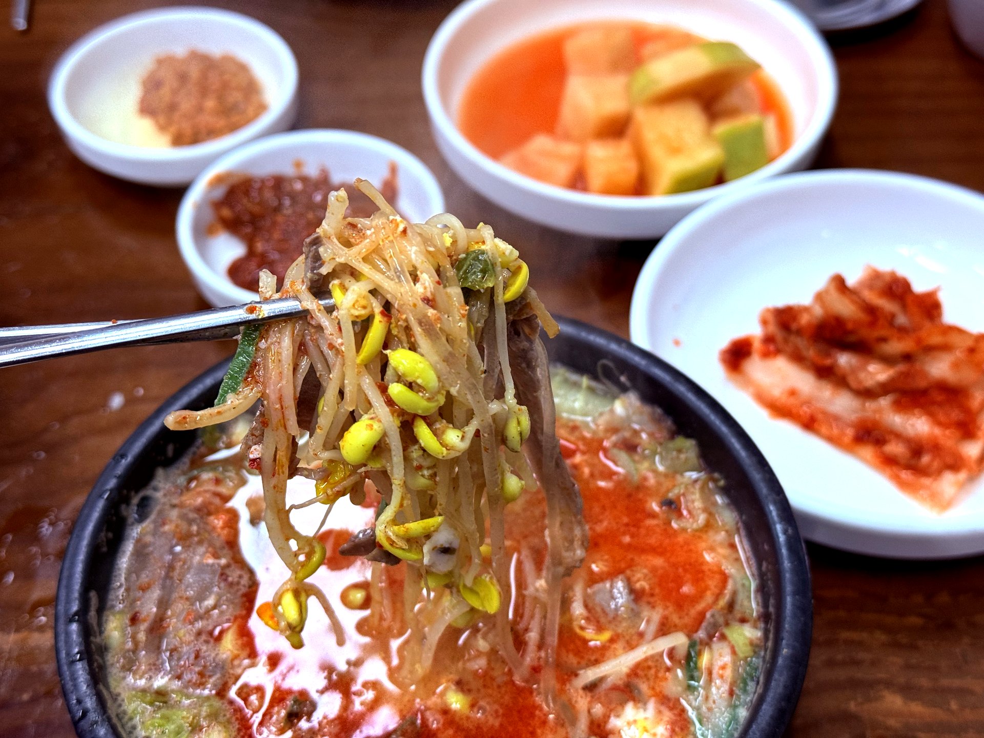 Bean sprouts lifted with chopsticks, banchan in frame