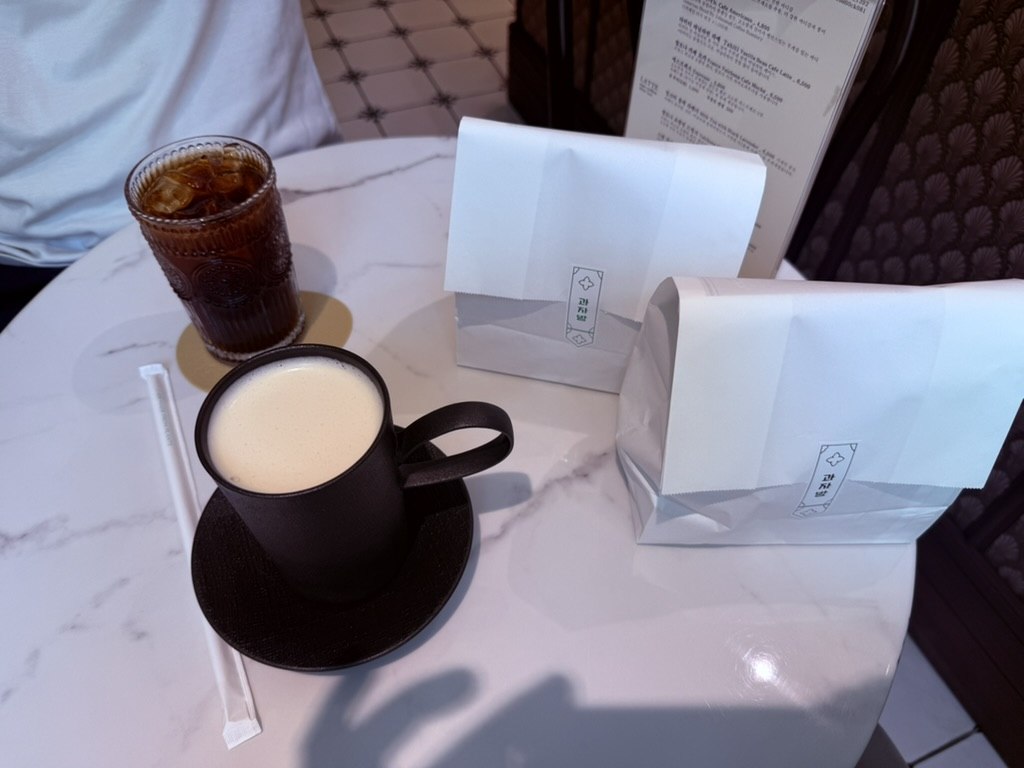 Full Gwajabang order - iced Americano in glass, milk tea in dark mug, two takeout paper bags with logo on white marble table