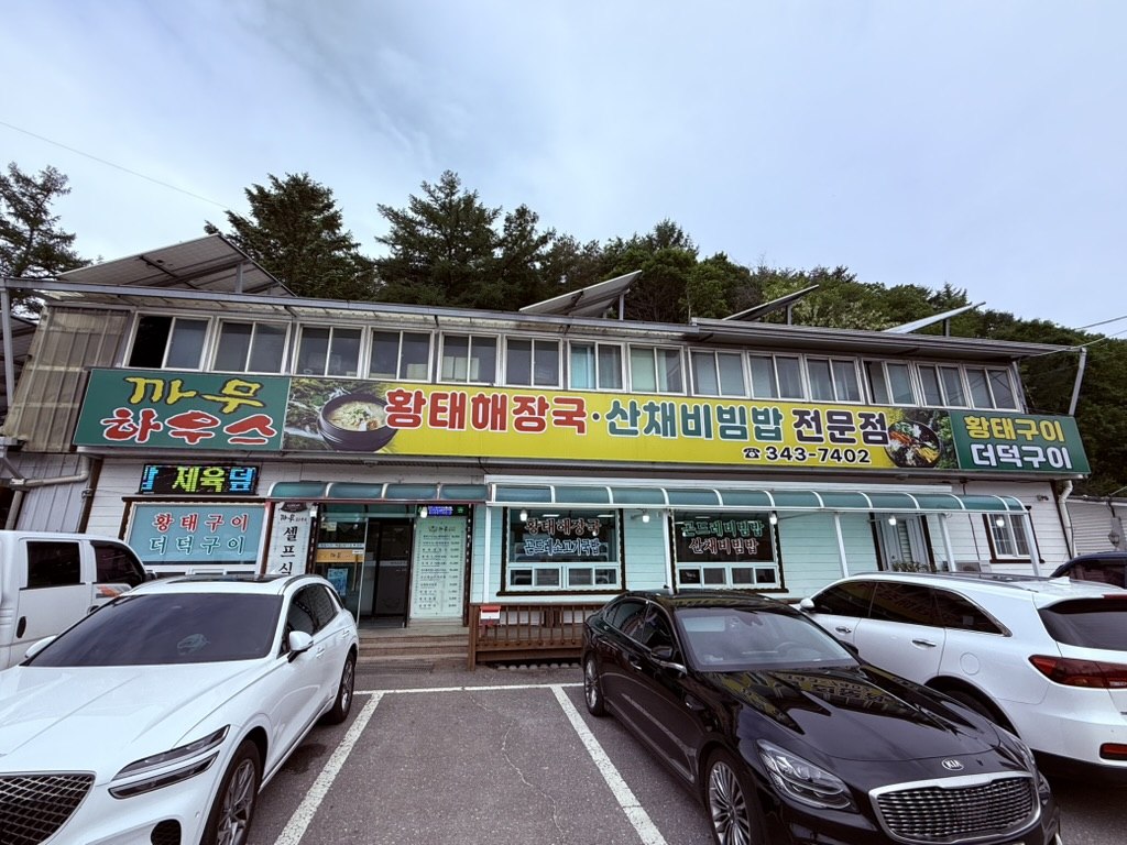 Kkamu House exterior - blue and orange Korean signage above single-story restaurant, mountain trees in background, parking lot with cars