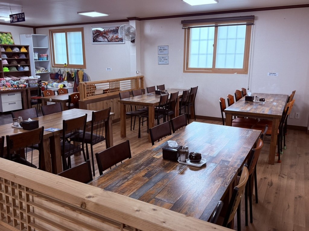 Kkamu House interior - wooden plank tables and chairs, big windows with mountain trees outside, simple craft shelf along one wall