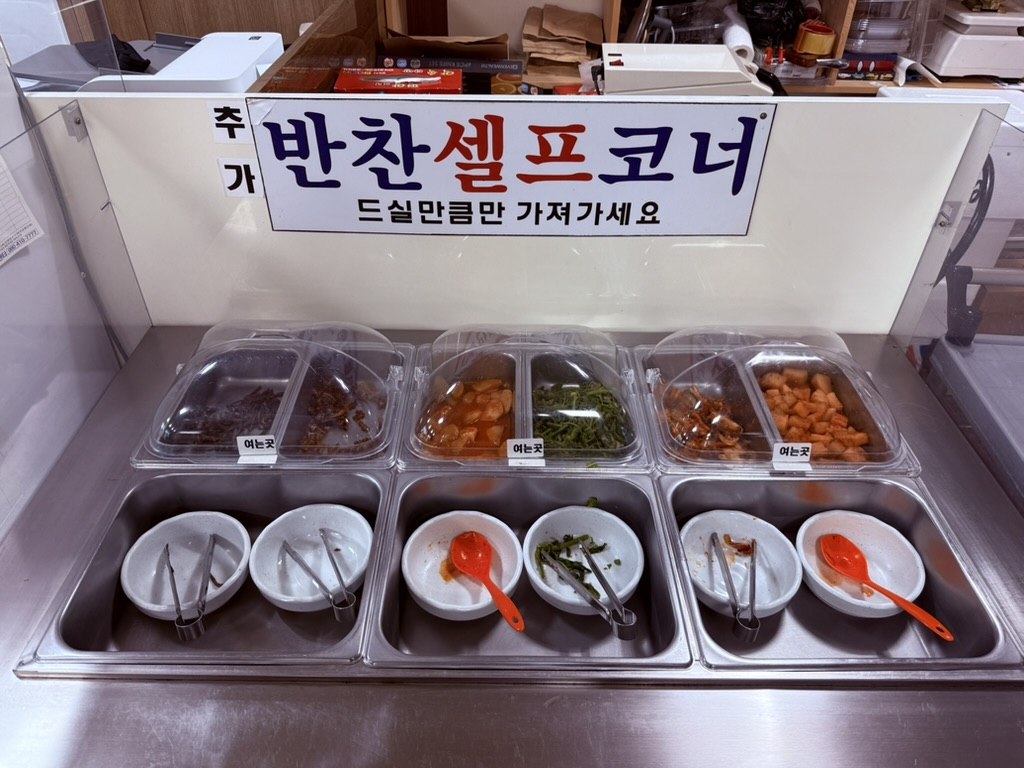 Kkamu House self-service banchan corner - large sign reading 반찬셀프코너 with six clear plastic bins of Korean side dishes, ice tongs and serving spoons in white bowls
