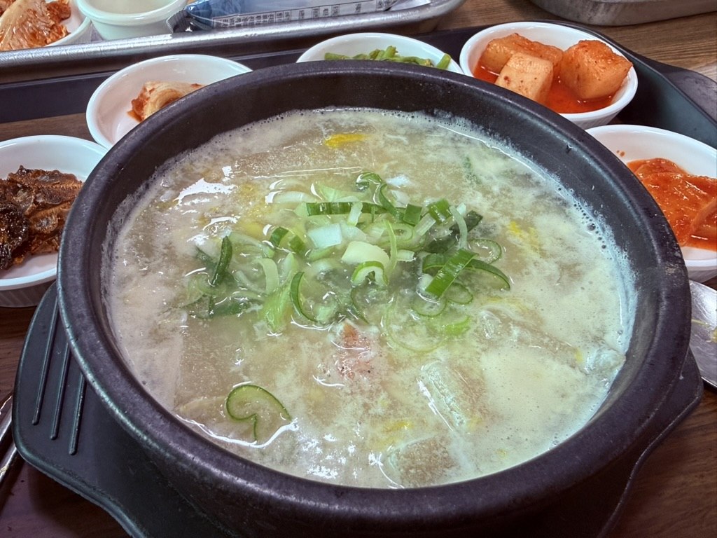 Kkamu House hwangtae haejangguk close-up - milky white pollock soup with sliced green scallions, in a black stone pot, surrounded by banchan