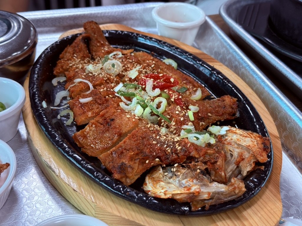Kkamu House grilled hwangtae pollock on a sizzling cast-iron platter - butterflied open, glazed in sweet-spicy red gochujang sauce, topped with toasted sesame seeds and green onion