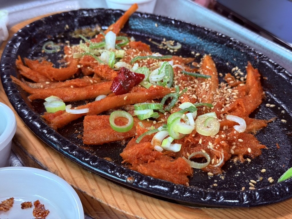 Kkamu House grilled deodeok root on cast-iron platter - long thin pieces of glazed deodeok in sweet-spicy red sauce, topped with green onion, sesame and dried chili
