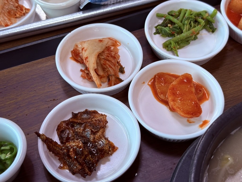 Three small white plates of Korean banchan at Kkamu House - white kimchi leaf, pickled radish in gochujang, and glazed anchovy and seaweed