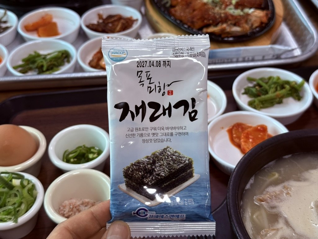 Packet of Korean roasted seaweed jaerae-gim held over the Kkamu House table - Korean text and ocean image on the package, banchan plates blurred in the background