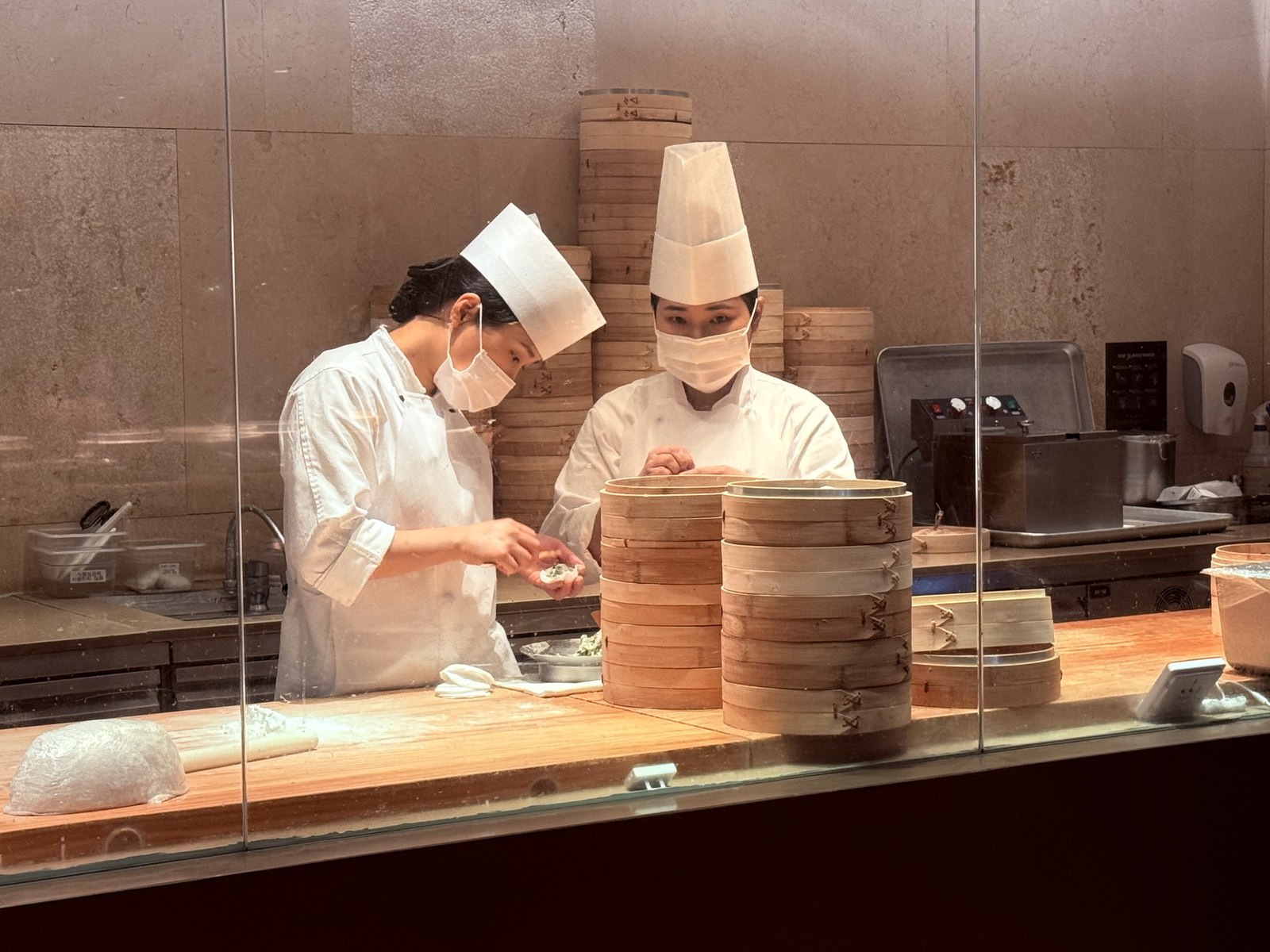 Two white-uniformed chefs hand-folding dim sum behind glass with stacked bamboo steamers