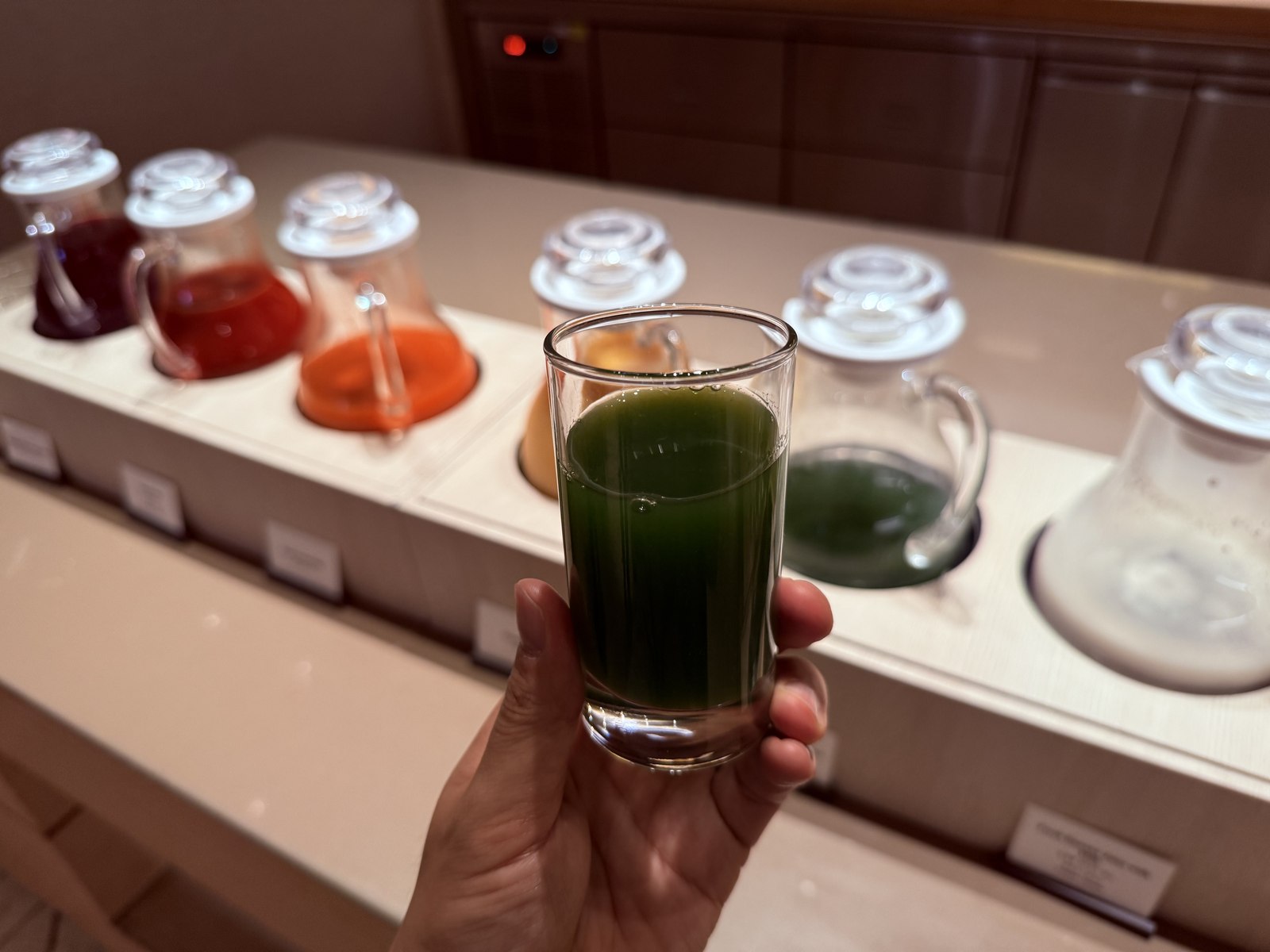 Hand holding clear glass of bright-green cold-pressed kale juice in front of juice carafes