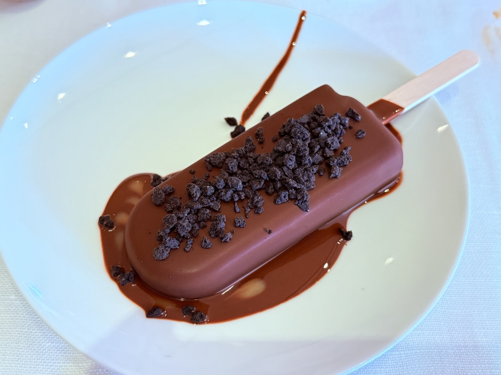 Chocolate-coated mango popsicle with raw chocolate dripping onto white plate, chocolate shards on top