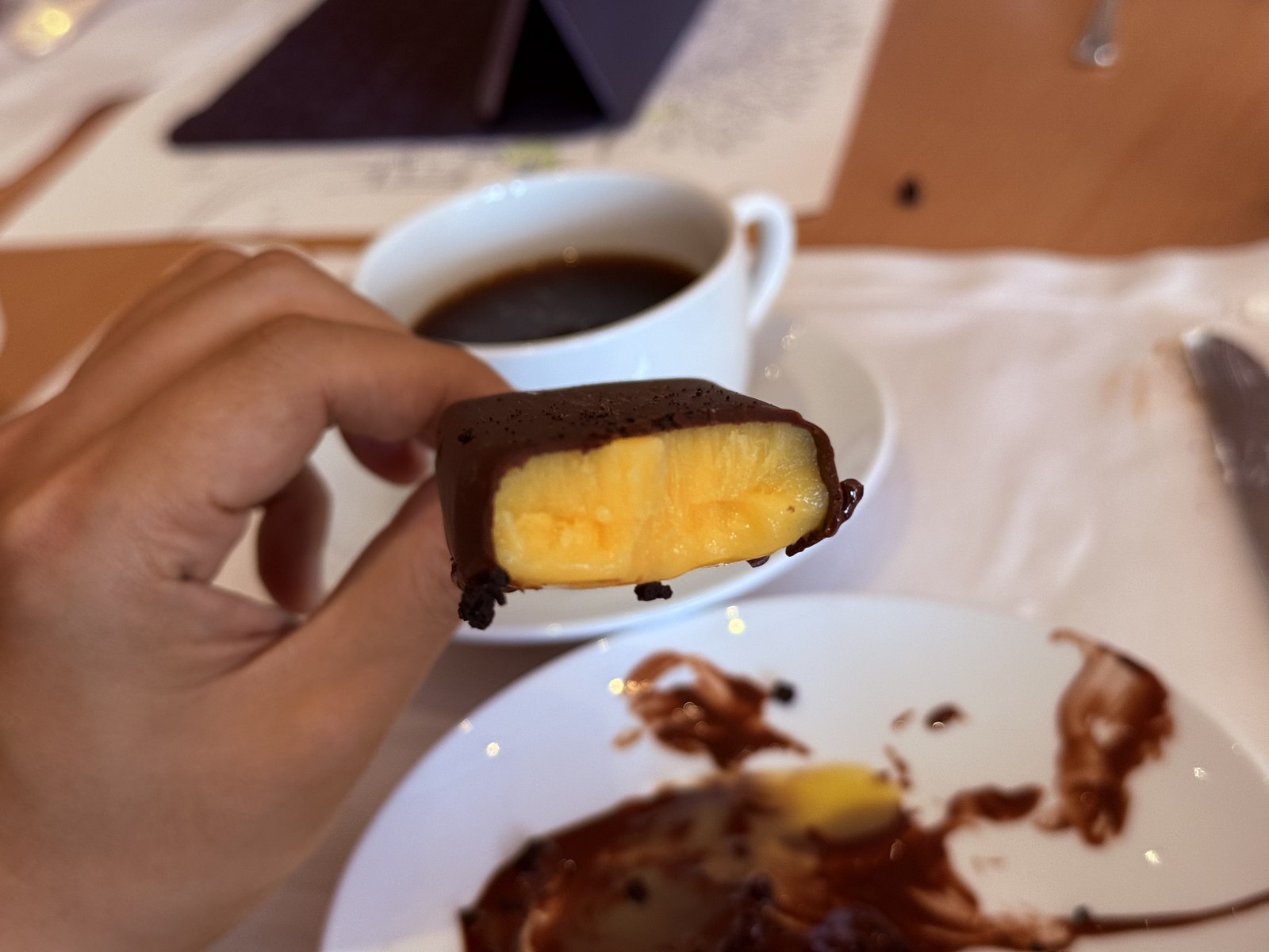 Hand holding chocolate-coated popsicle with mango interior visible, cup of black coffee in background