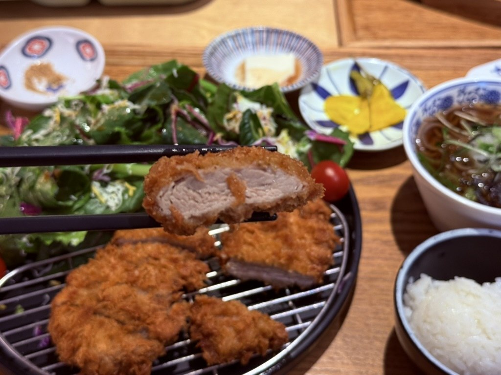 Chopsticks lifting a piece of Bueongi tenderloin donkatsu showing the crisp golden panko exterior and pink juicy pork tenderloin interior - cross-section close-up with the full plate and salad in background