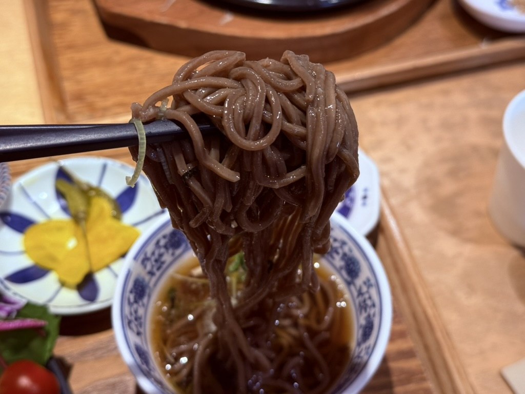 Action shot of Bueongi cold buckwheat soba mini side - chopsticks lifting brown buckwheat noodles from a blue-and-white porcelain bowl of cold dashi broth, with the donkatsu set and pickled radish in the background