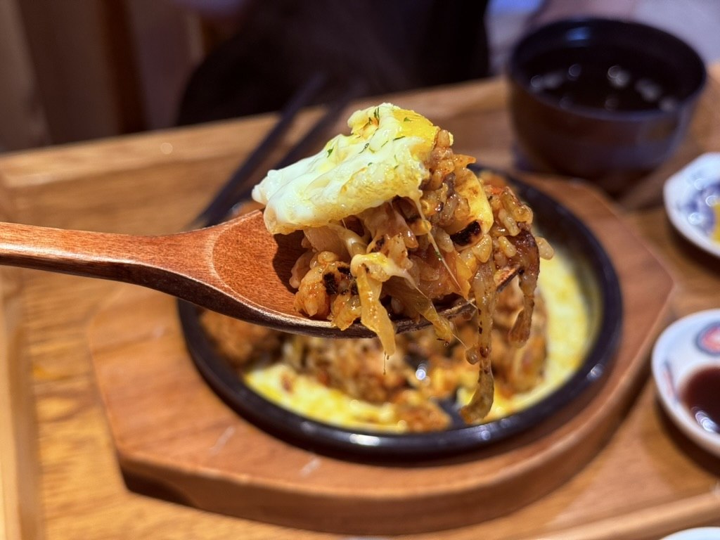 Extreme close-up of a wooden spoon holding a perfect bite of Bueongi kimchi cheese fried rice with melted mozzarella, kimchi-stained rice, sunny egg yolk and donkatsu - steam visible, the bowl half-eaten in the background