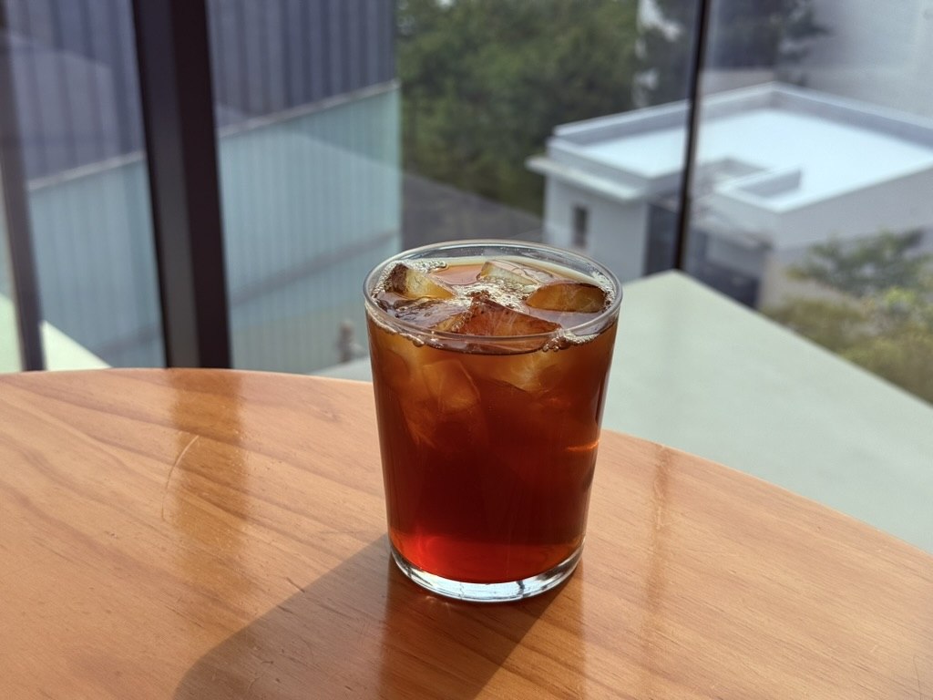 Derban Berlin Busan iced Brazil Daterra Reserve Low Caf drip coffee - clean clear glass of cold-brewed pour-over coffee in amber-red color with ice cubes, sitting on a circular wooden side table backlit by the cafe's large windows
