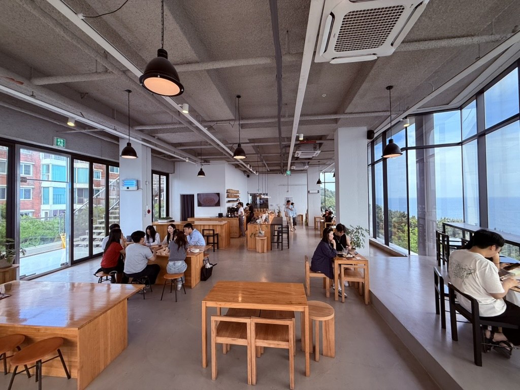 Derban Berlin Busan interior wide view - modern industrial concrete cafe with exposed pipes, large floor-to-ceiling windows facing the ocean on the right side, raw wooden tables and stools, customers seated and working, drip coffee bar visible in the background