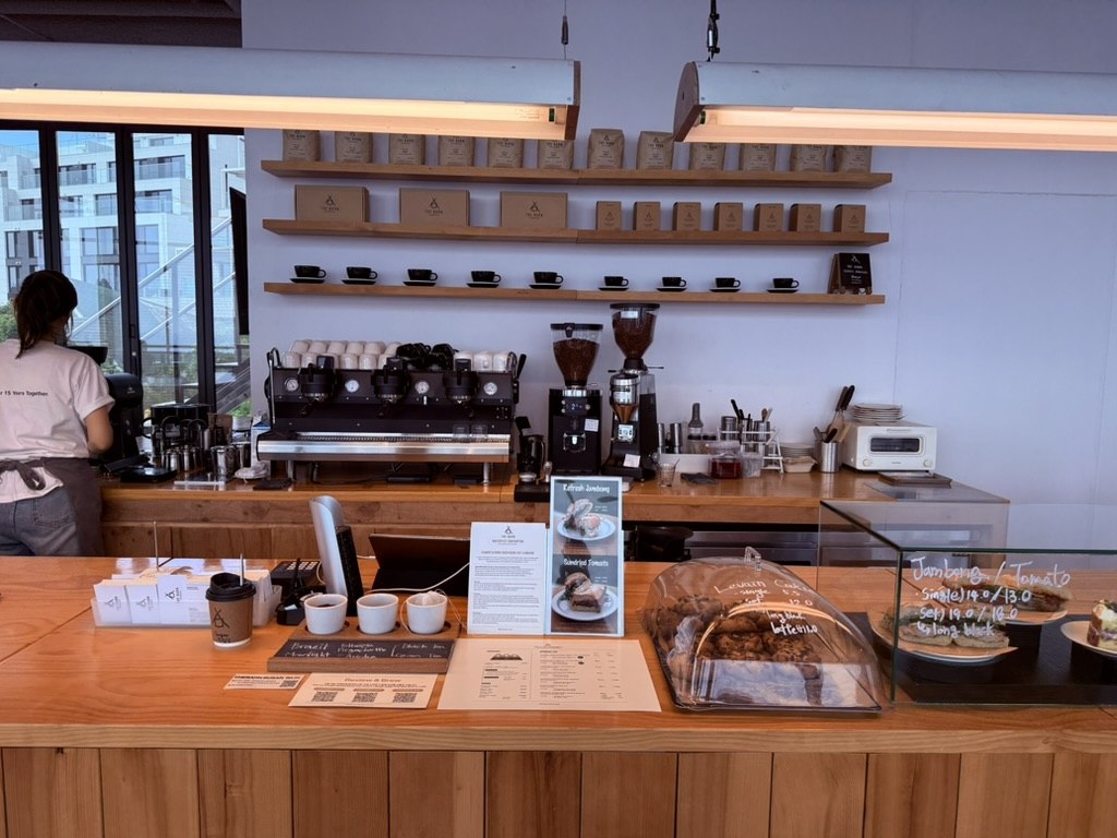 Derban Berlin Busan barista bar - clean modern setup with two-group espresso machine, conical coffee grinders, hand-built shelf of black ceramic espresso cups, kraft-paper coffee bags lined up on shelves above with THE BARN logo, pastry case visible in foreground with hand-written menu items