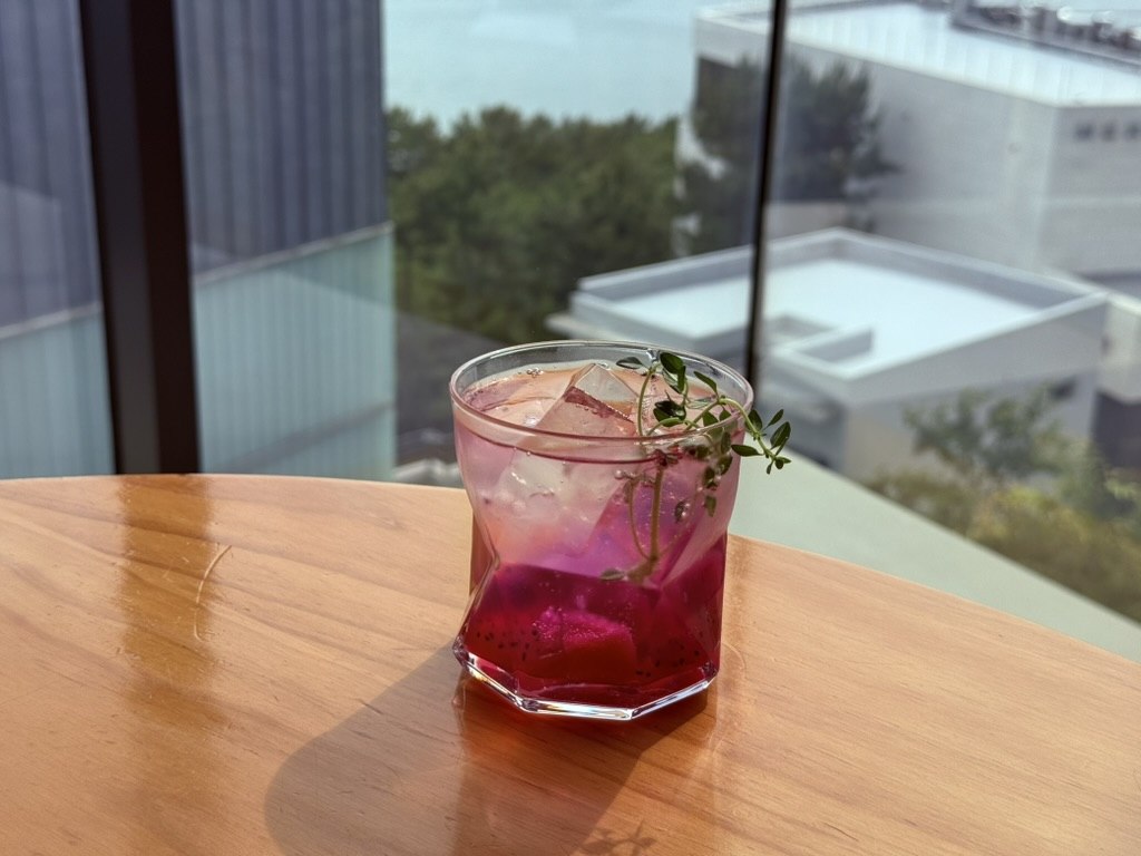 Derban Berlin Busan seasonal ade close-up - vivid magenta-pink dragonfruit ade in a faceted glass with ice cubes, fresh dragonfruit chunks visible at the bottom, a sprig of fresh thyme floating on top, on a wooden table with the floor-to-ceiling Haeundae view in the background