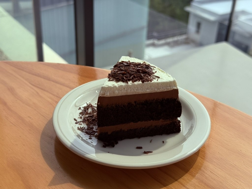 Derban Berlin Busan Choco Fudge cake slice - thick triangular slice of chocolate cake with two dark chocolate cake layers, dark chocolate ganache filling, a white cream top layer, finished with dark chocolate shavings, on a white ceramic plate on a wooden table