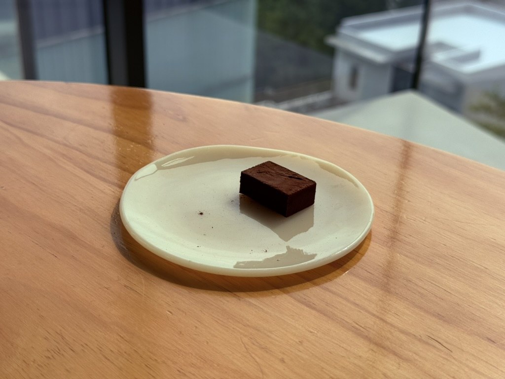 Single chocolate truffle on a cream-colored ceramic plate at Derban Berlin Busan - small dark chocolate square dusted with cocoa powder, minimalist composition on a wooden table with windows blurred in the background