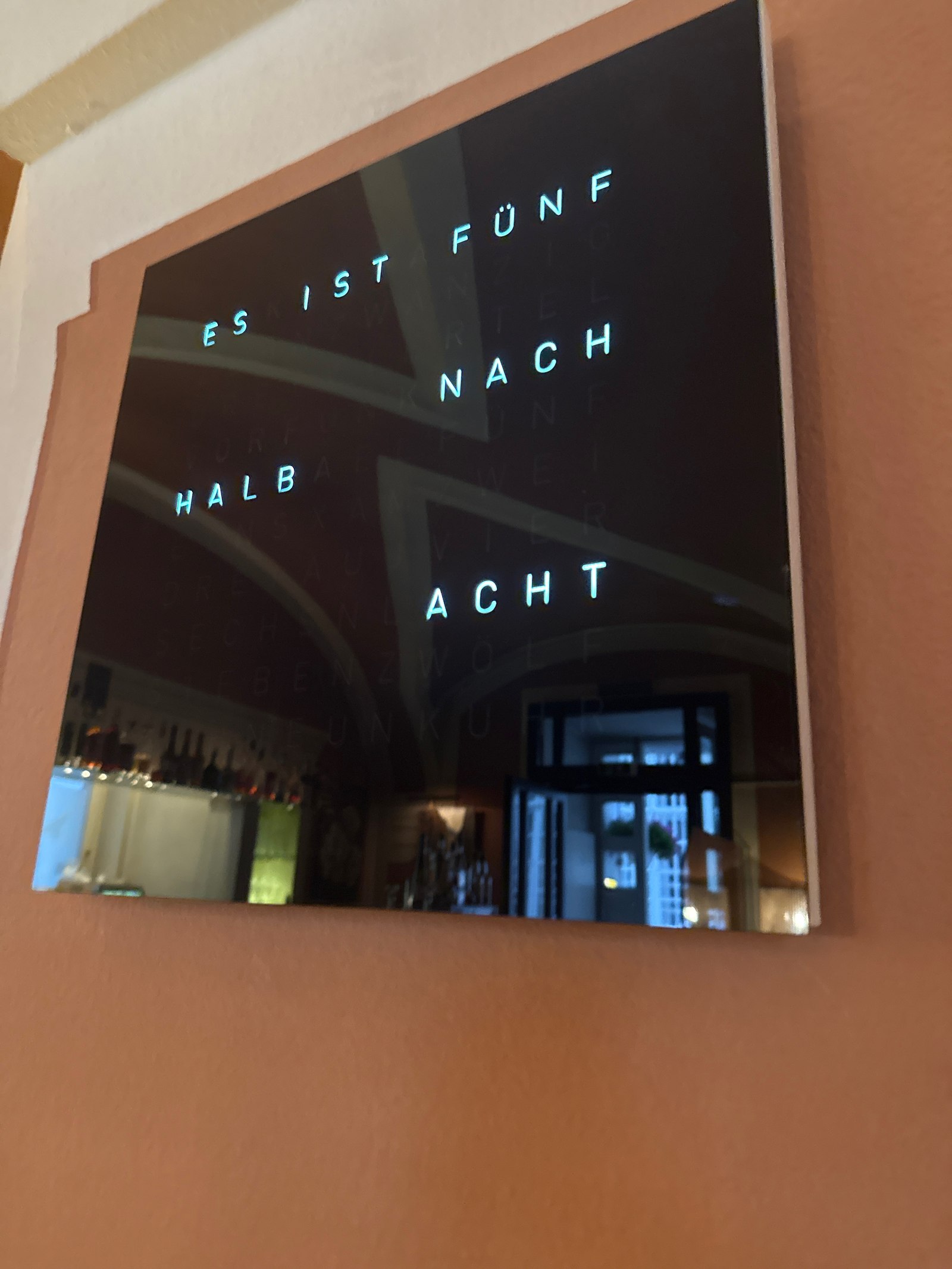 QlockTwo word clock at Restaurant Duchardt - square electronic word clock mounted on a salmon-orange wall, lit cyan-blue text reading ES IST FÜNF NACH HALB ACHT (German for 7:35), the dark mirror surface reflecting the restaurant bar and dining room behind