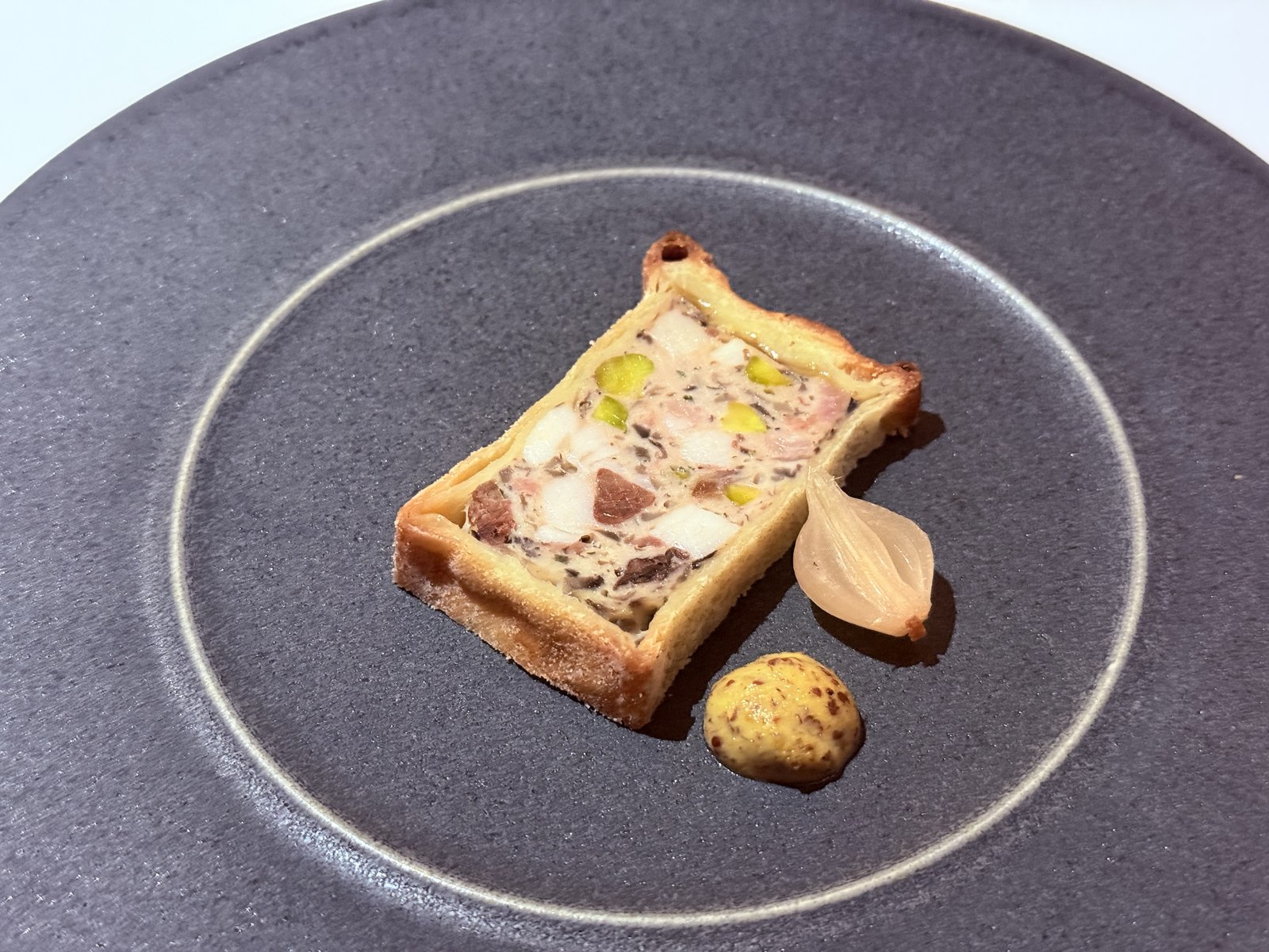 Restaurant Duchardt Paté en Croûte first course - rectangular slice of French pastry-crust terrine showing visible cubes of pork, chicken liver, green pistachios and white fat marbling, accompanied by a single pickled shallot and a small mound of grain mustard on a dark grey stoneware plate