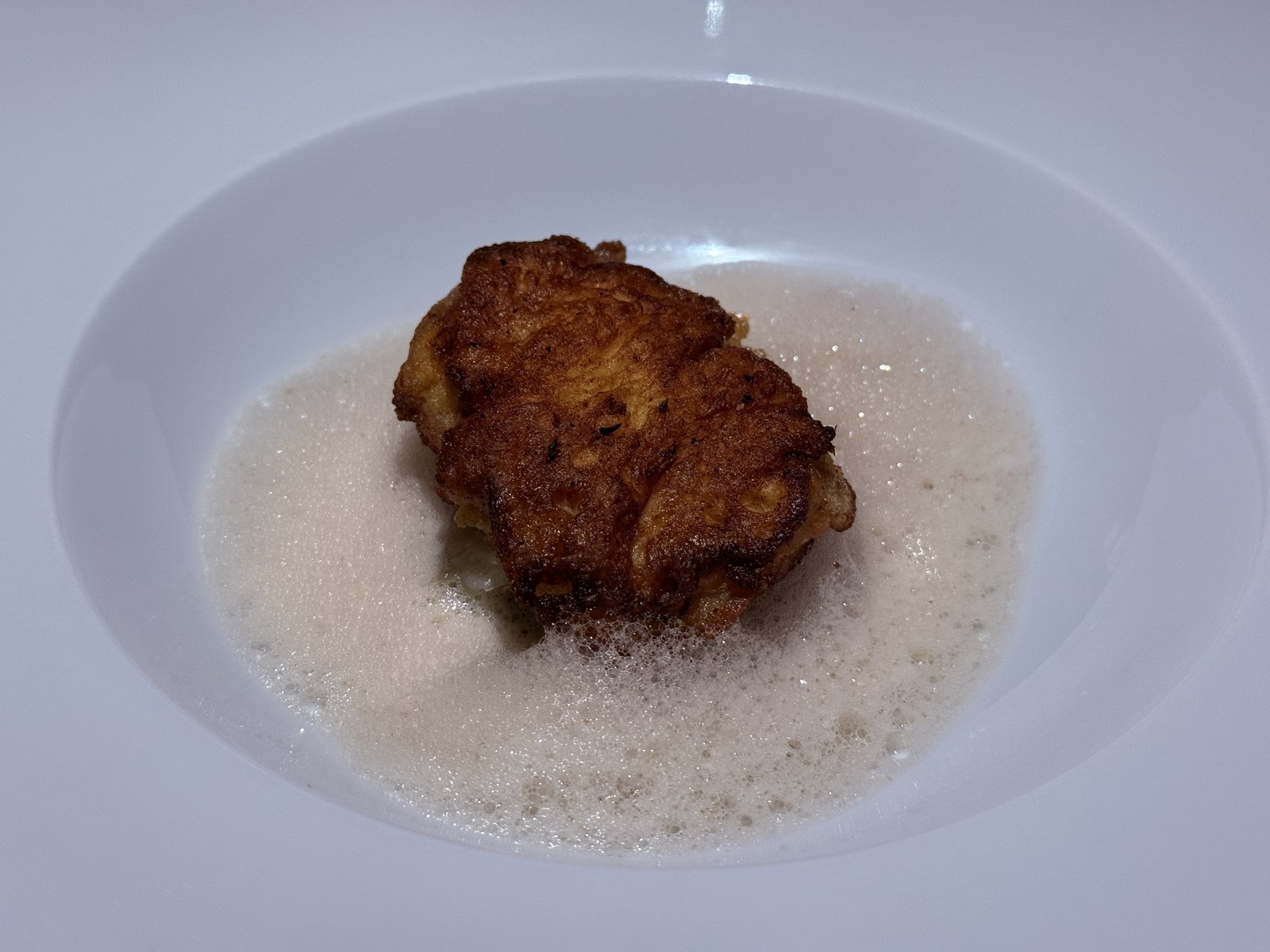 Restaurant Duchardt Monkfish picatta third course - a golden egg-battered fried monkfish piece sitting in a foamy cream-colored Japanese dashi broth with shaved fennel, in a wide white deep bowl, the foam visible across the surface