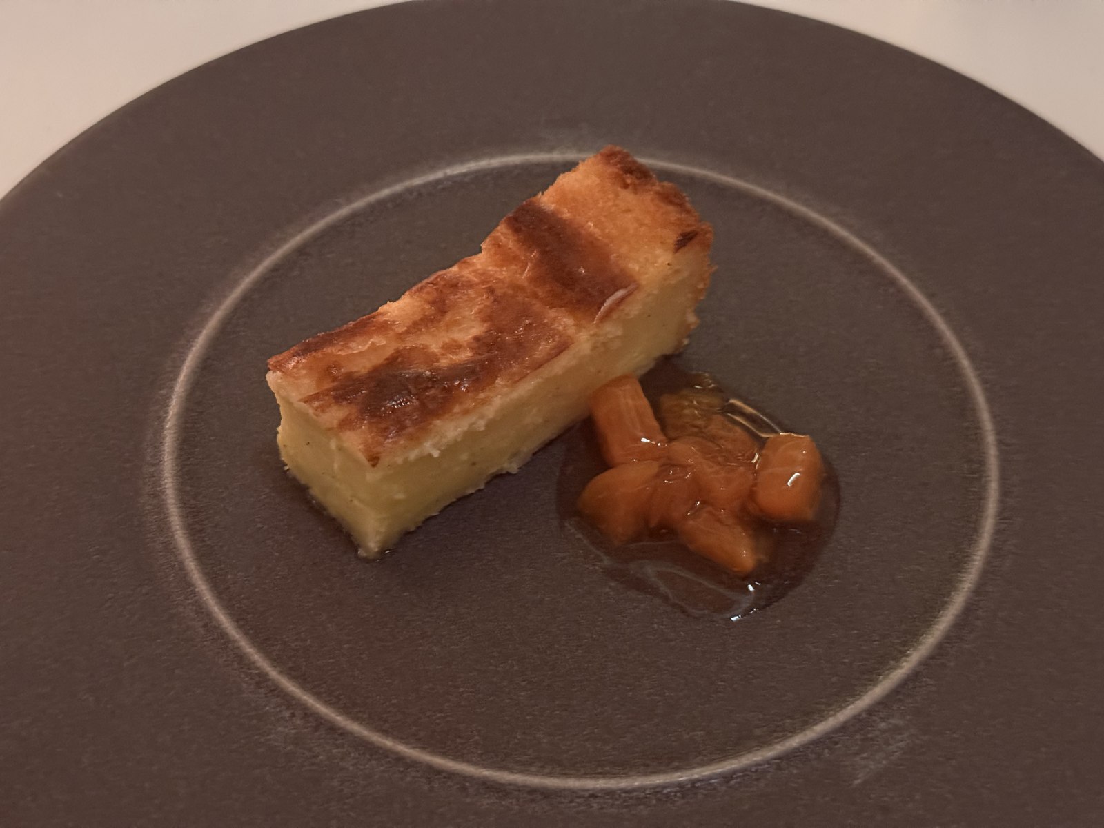 Restaurant Duchardt Gâteau Basque fifth course dessert - a rectangular slice of golden-crusted French Basque cake with a darker caramelized top, served with a small mound of glossy poached apricot compote on a dark grey stoneware plate