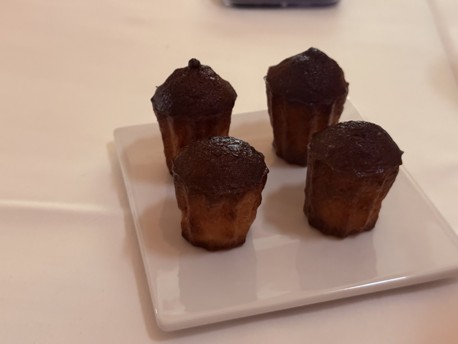 Restaurant Duchardt closing course petit fours - four miniature canelé-style dark-caramelized French pastries with glossy tops and pale fluted bases, arranged in a square pattern on a white ceramic plate