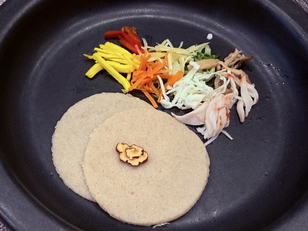 Naedang buckwheat crepes with nine delicacies - two pale beige round buckwheat crepes on a black ceramic plate, with a small pile of julienned colorful fillings including yellow squash, carrot, red pepper, white cabbage, crab meat and mushrooms beside them