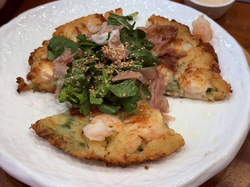 Naedang shrimp jeonyuhwa with jamon and rocket salad - large round golden Korean pan-fried shrimp pancake quarters with pink jamon ribbons, fresh rocket leaves and sesame seeds piled in the center on a wide white plate