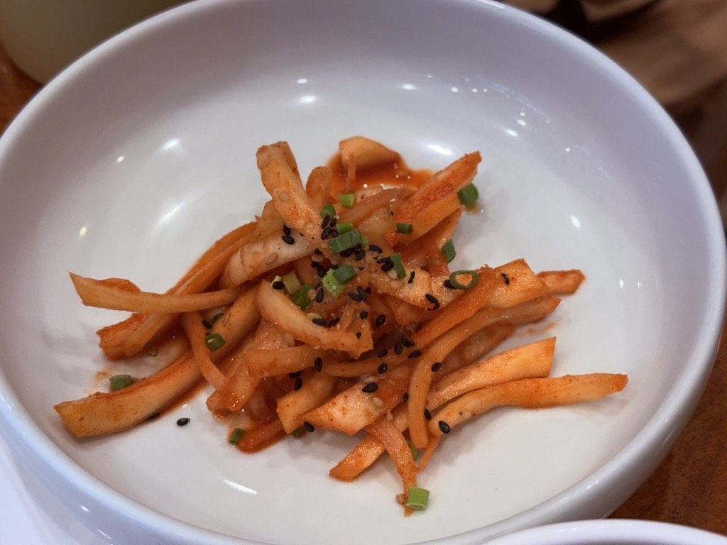Naedang mu saengchae spicy Korean radish salad close-up - julienned daikon strips dressed in red gochugaru chili flakes with chopped scallion and black sesame seeds in a small white round bowl