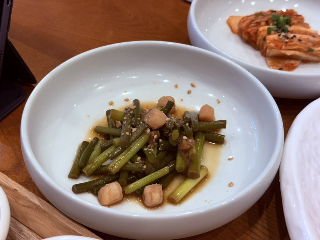 Naedang garlic stems with daikon cubes - bright green chopped garlic-stem batons with small cubes of pickled daikon radish in a soy-vinegar dressing with sesame seeds, in a small white bowl, with kimchi visible in the background