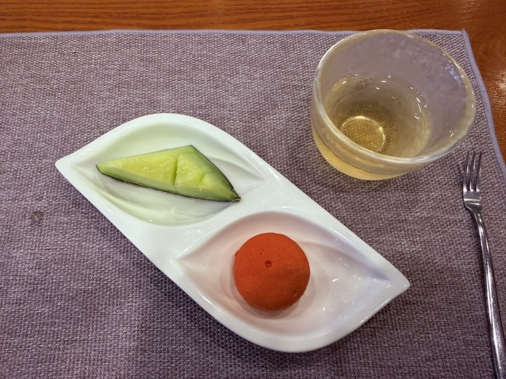 Naedang traditional Korean tea and dessert close-up - white rectangular ceramic plate with a green wedge of fresh melon and a small round red sweet-rice cake (경단 gyeongdan), with a small clear glass cup of warm yellow chrysanthemum tea on a lavender linen tablecloth