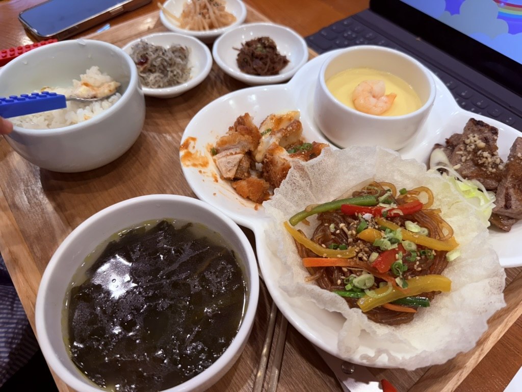Naedang kids meal set on a wooden tray - bowl of white rice with a colorful blue Lego-themed spoon, japchae glass noodles with bell peppers in a crispy fried rice paper cup, mini Korean donkkaseu pork cutlets, seaweed soup, steamed egg custard with shrimp on top, small tteokgalbi, and Korean banchan dishes - clearly designed for children