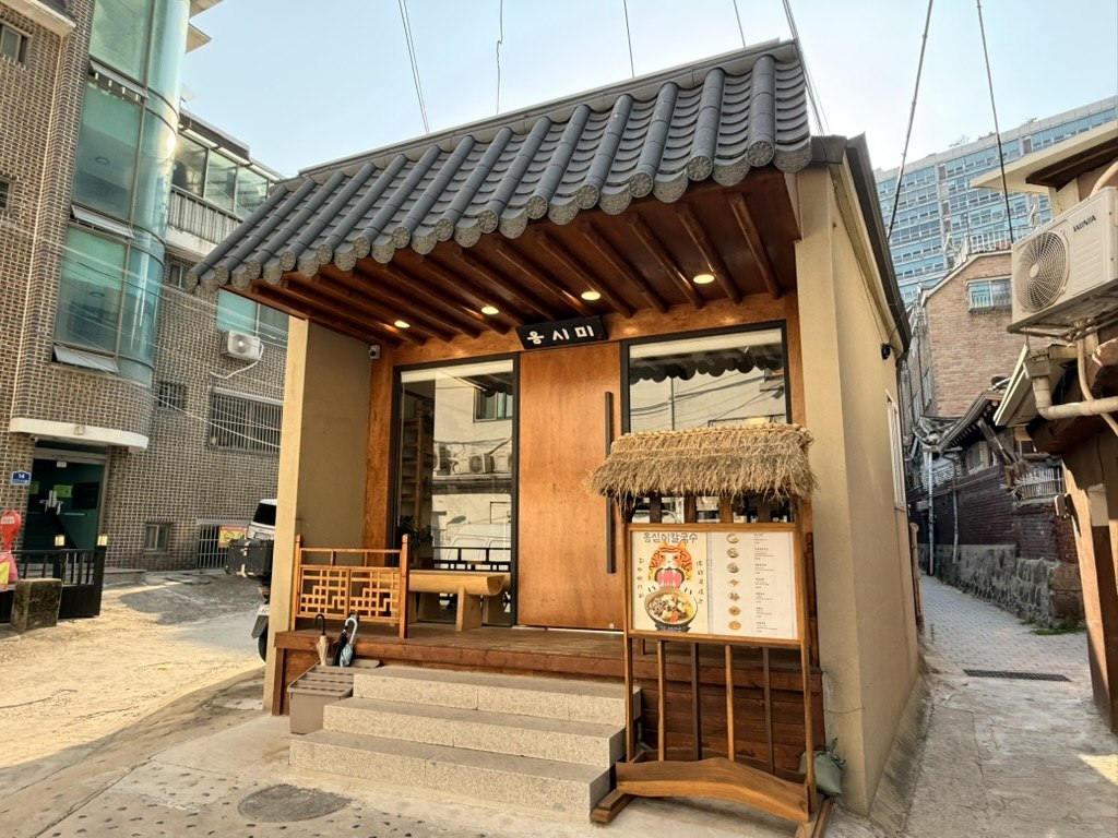 Ongsimi Seoul Station hanok exterior - small standalone single-story Korean traditional house with grey tile roof, wooden facade, small black plaque reading 옹시미 (Ongsimi), wooden steps up to glass door, and a small thatched-roof menu stand displaying the famous Korean potato dumpling soup photo on the right, in a quiet Seoul side street near Seoul Station