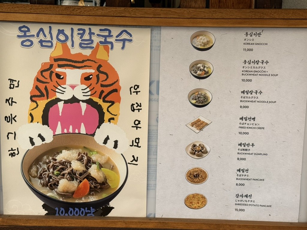 Ongsimi Seoul Station menu board - a Korean tiger poster on the left advertising 옹심이칼국수 (Korean Potato Gnocchi Soup) at 10000 won, and on the right a price list showing all 8 dishes with Korean and English names: Korean Gnocchi, Buckwheat Noodle Soup, Fried Kimchi Crepe, Buckwheat Dumpling, Buckwheat Pancake, Shredded Potato Pancake