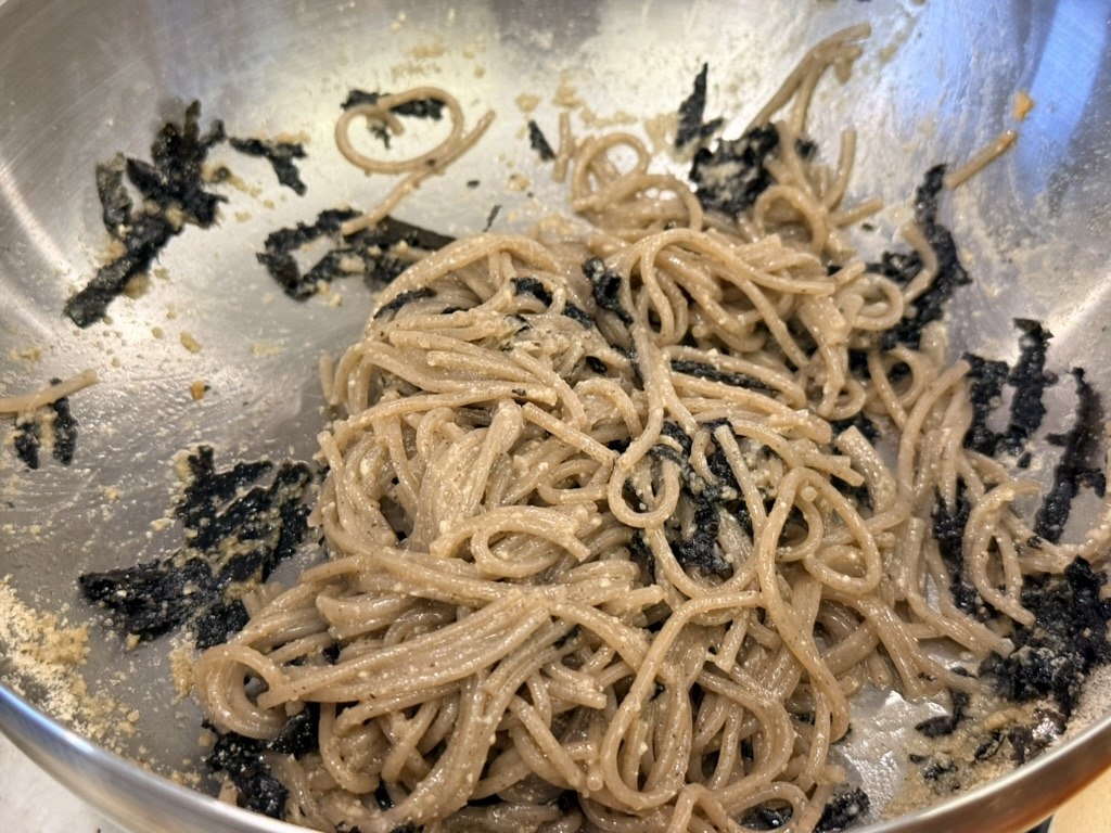 Ongsimi Seoul Station 들기름 막국수 (perilla-oil buckwheat noodles) - a stainless steel bowl of cold brown buckwheat noodles glistening with perilla oil and tossed with shredded toasted seaweed (gim) flakes, the noodles partially eaten with the seaweed clinging to the sides of the bowl