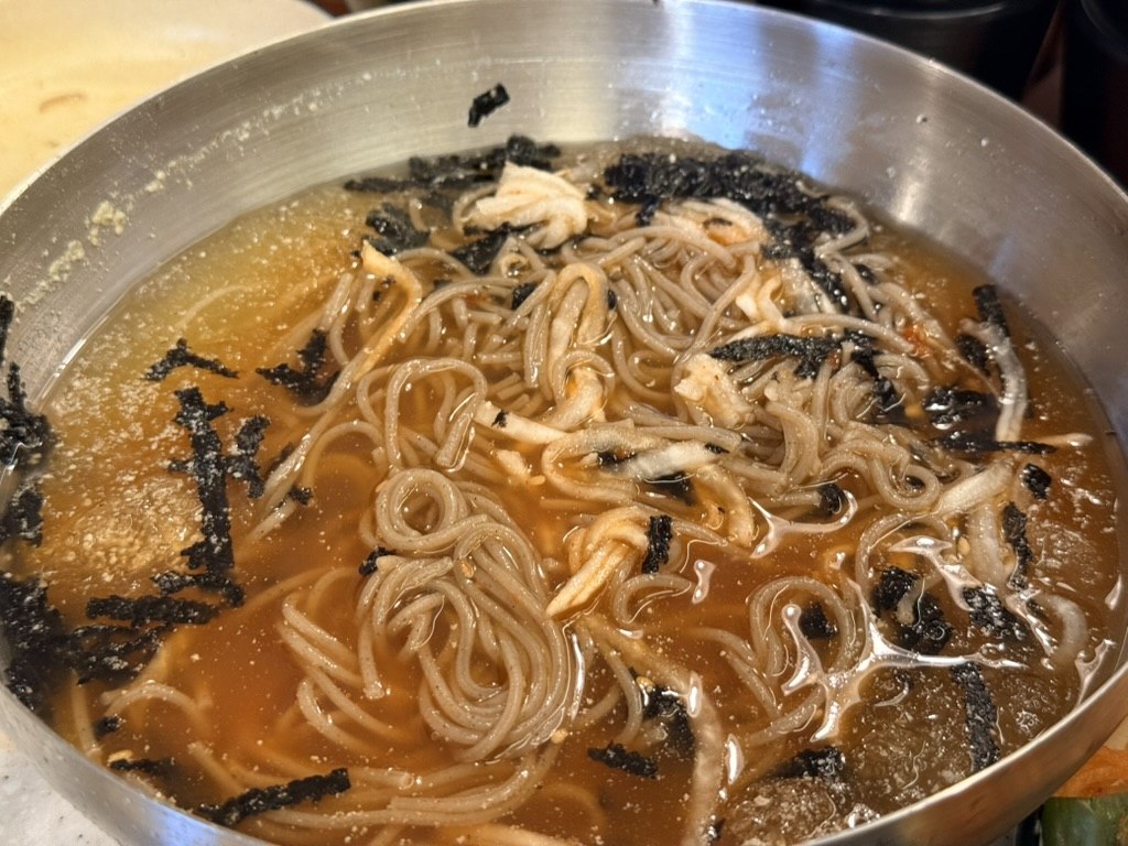 Ongsimi Seoul Station 물 막국수 (cold buckwheat noodles in dongchimi broth) - large stainless steel bowl of brown buckwheat noodles submerged in a clear tangy radish-water broth with shredded toasted seaweed flakes scattered on top and a hint of young radish leaves visible underneath