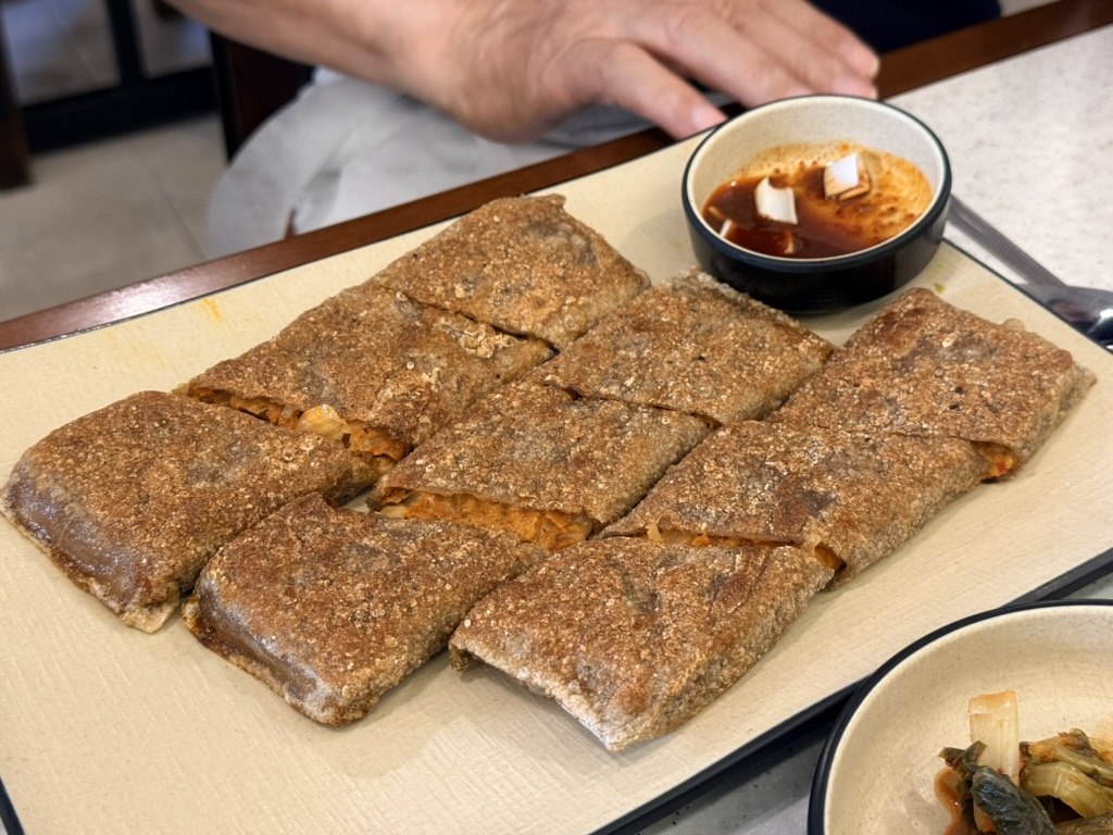 Ongsimi Seoul Station 메밀전병 (buckwheat-kimchi crepe) - eight rectangular slices of thin brown buckwheat crepe rolled around stir-fried aged kimchi filling, dusted with toasted barley powder, arranged in two rows on a beige rectangular tray with a small bowl of red gochujang dipping sauce in the upper right corner