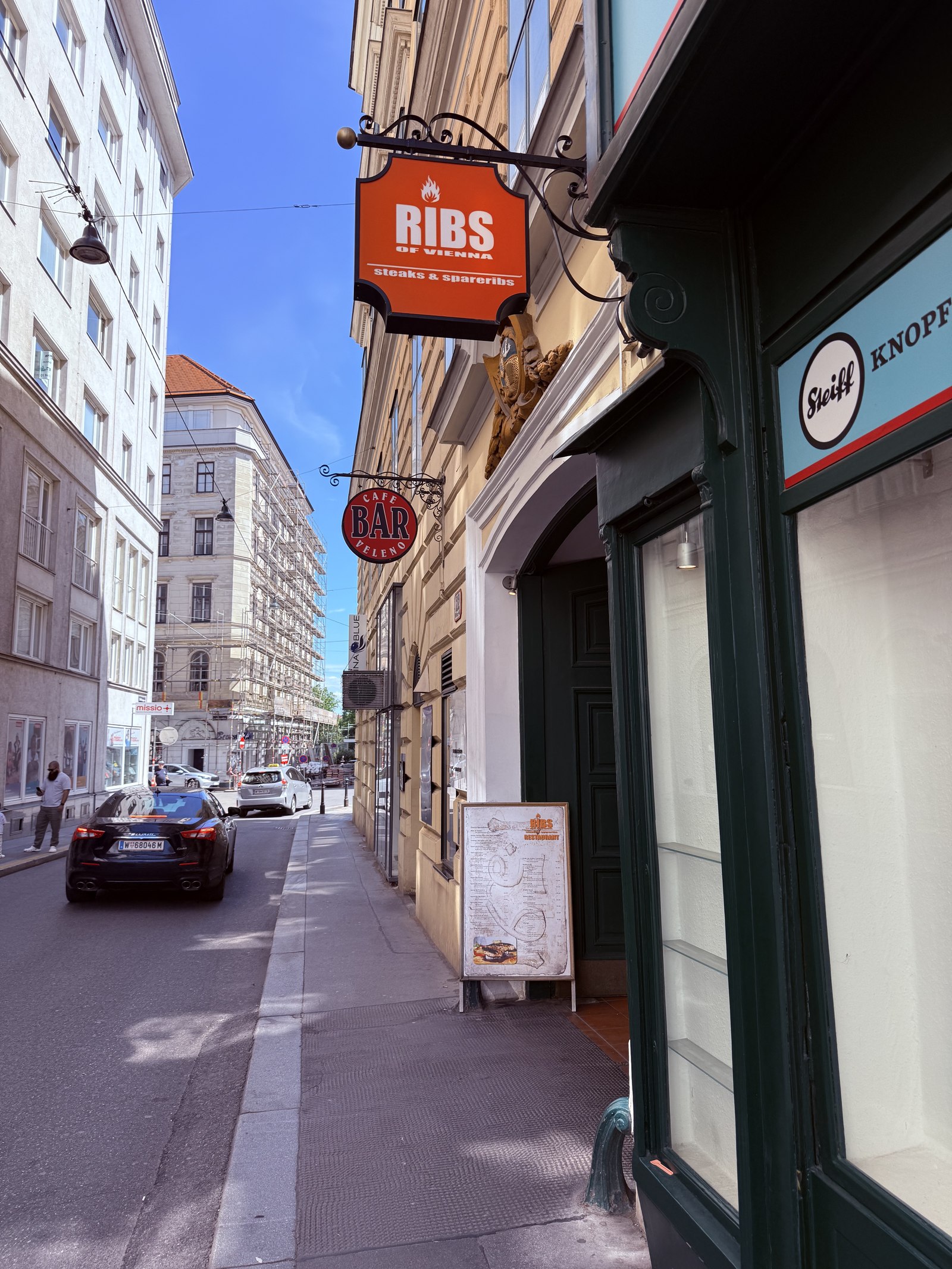 Ribs of Vienna exterior - hexagonal hanging blade sign reading RIBS in white on red, with TRADE & RESTAURANT underneath, a red BAR neon sign, and an awning over the door on a Vienna Inner Stadt street, classic European buildings visible
