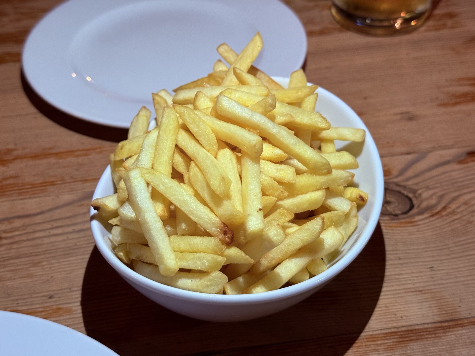 Bowl of yellow shoestring French fries piled high in a white ceramic bowl on a worn wooden table at Ribs of Vienna, with a half-empty beer glass and edge of an empty plate visible