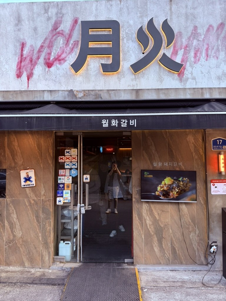 Wolhwa Galbi exterior in Mullae - concrete facade with large painted 월화 sign in pink graffiti calligraphy, a flame icon, 월화갈비 Korean text below the awning, glass door entrance with menu posters, modern industrial Mullae street setting