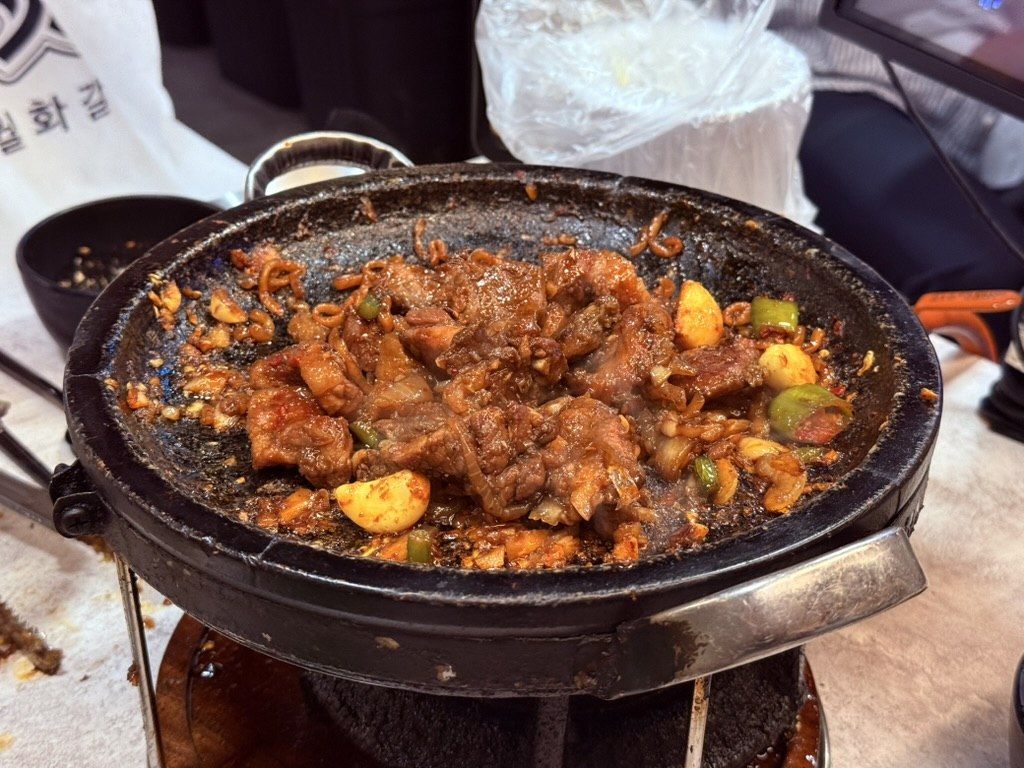 Wolhwa Galbi 마늘폭탄 garlic-bomb pork galbi - smaller pieces of marinated pork mixed with whole roasted garlic cloves and green chili pepper pieces on the stone grill, with visible 월화갈비 signage in the background