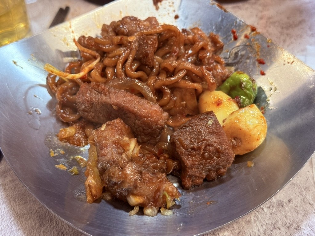 Wolhwa Galbi 파김치 짜파구리 after-dish - dark jjajang-style instant noodles in a metal bowl with chunks of grilled pork galbi, pickled scallions, green chili, and a touch of sauce, the Parasite-movie chapaguri reference dish