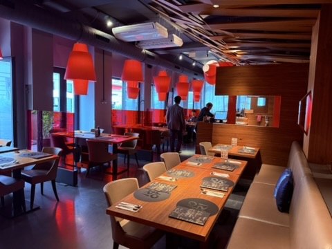 YORI Vienna interior - modern dining room with wooden tables and beige bench seating, warm red pendant lamps hanging over each table, wood-clad ceiling with diagonal beams, glass partitions, two diners at a booth in the background