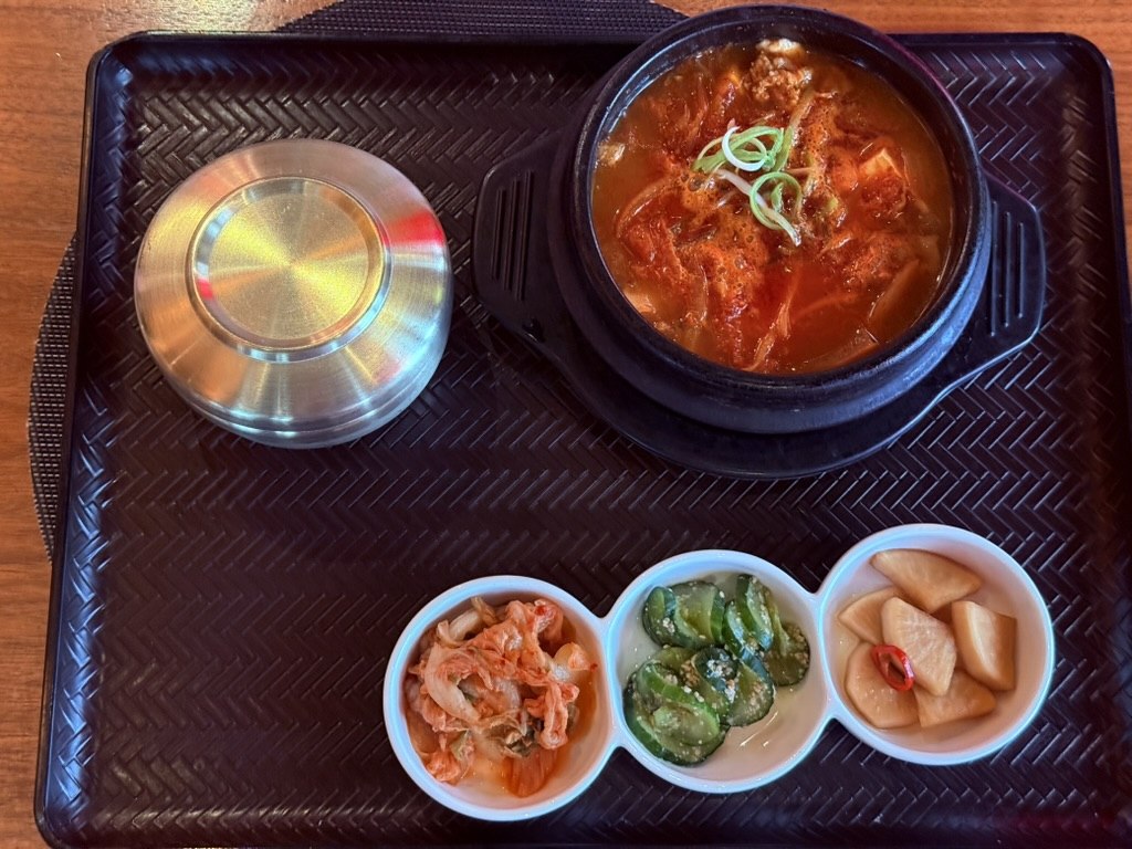 YORI Vienna kimchi jjigae set - bubbling red stone pot of kimchi stew with tofu cubes, kimchi pieces and scallion on top, beside a covered stainless rice bowl with lid, and a 3-bowl banchan tray with cabbage kimchi, sesame cucumbers and pickled daikon, on a black woven tray